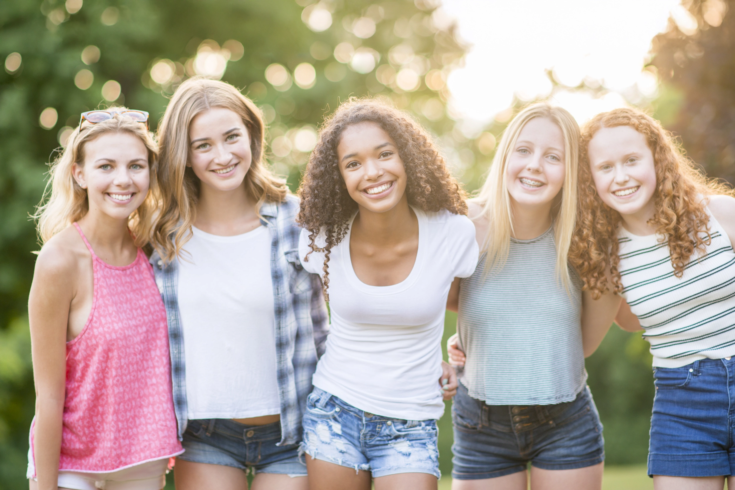 Gruppo di ragazze teenagers, sorridenti, vestite casual, in ambiente esterno. Sullo sfondo ci sono degli alberi.